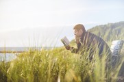 man sitting on bench reading a book. Bench surrounded by tall grasses, sun shining, portion of lake visible in background